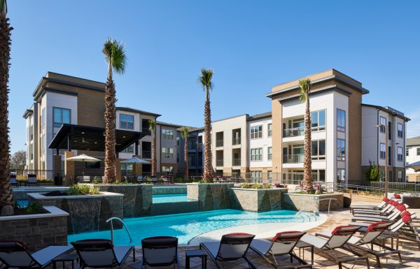 Sparkling, blue pool and expansive sundeck with ample lounge chairs, private cabana, and the clubhouse and apartment buildings in the background