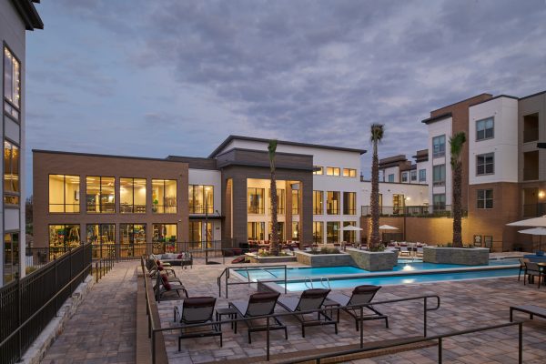 Sparkling, blue pool and expansive sundeck with ample lounge chairs and the clubhouse and apartment buildings in illuminated the background at dusk